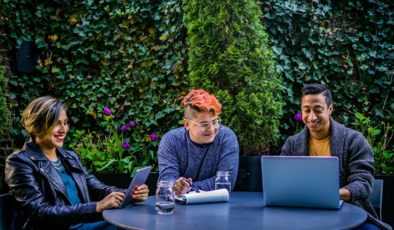 Three diverse adults engaging in an outdoor meeting with laptops and tablets in a garden.