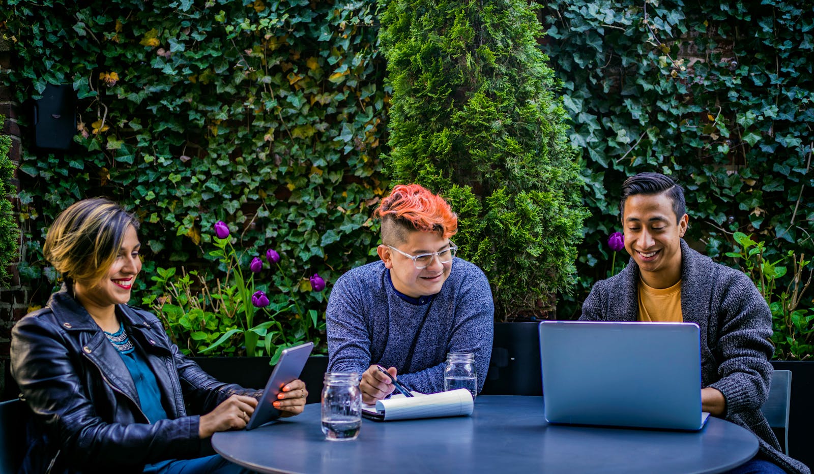 Three diverse adults engaging in an outdoor meeting with laptops and tablets in a garden.